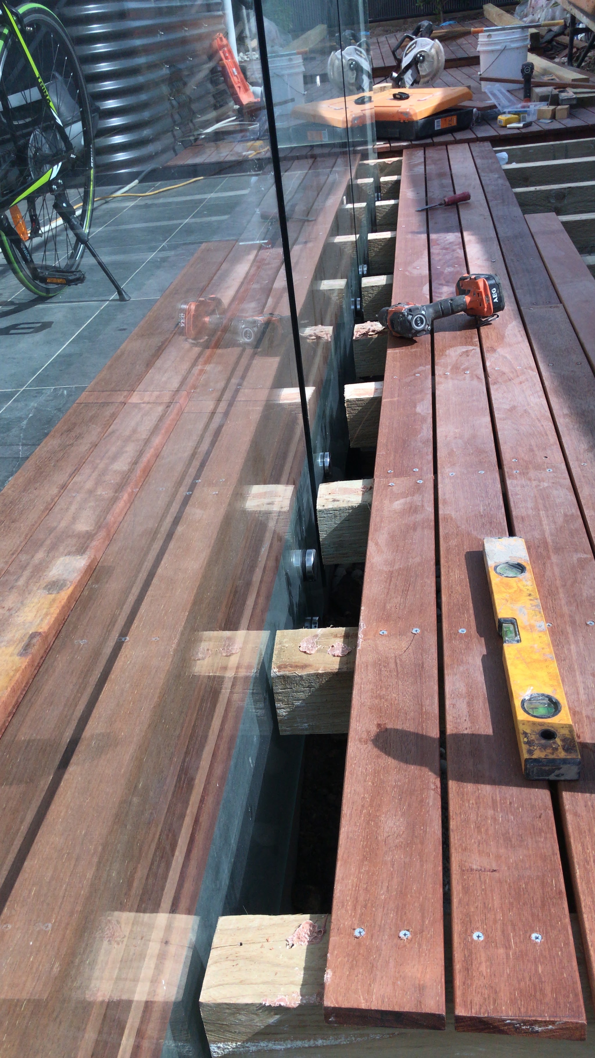 Hardwood deck boards being laid beside a frameless glass balustrade, with a drill, spirit level and saws on the boards